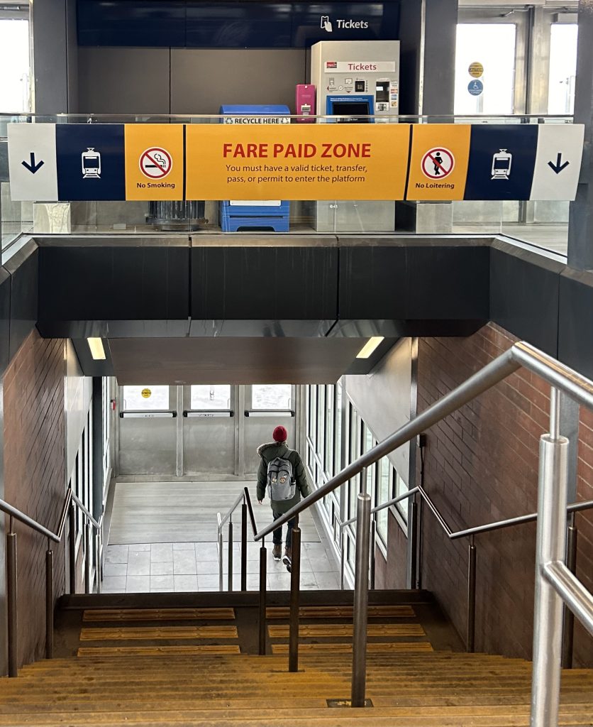 A man walks down the stairs at University Station along the Red Line of the C-Train in Calgary on Monday, March 9, 2026. The signage that hangs above the stairs is a reminder about paying the fare, with many machines to purchase and verify tickets surrounding the upper platform.